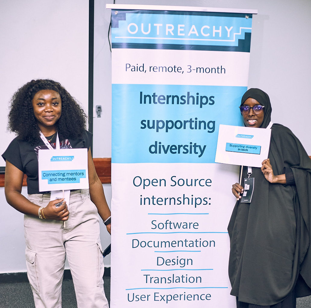 Two black people are standing by an Outreachy banner. On the left, Omotola Omotayo, holds a sign that reads 'Outreachy - connecting mentors and mentees'. The Outreachy banner says 'Outreachy - Paid remote 3-month internships supporting diversity. Open source internships: software, documentation, design, translation, user experience.' Photo is cc-by 4.0 Outreachy